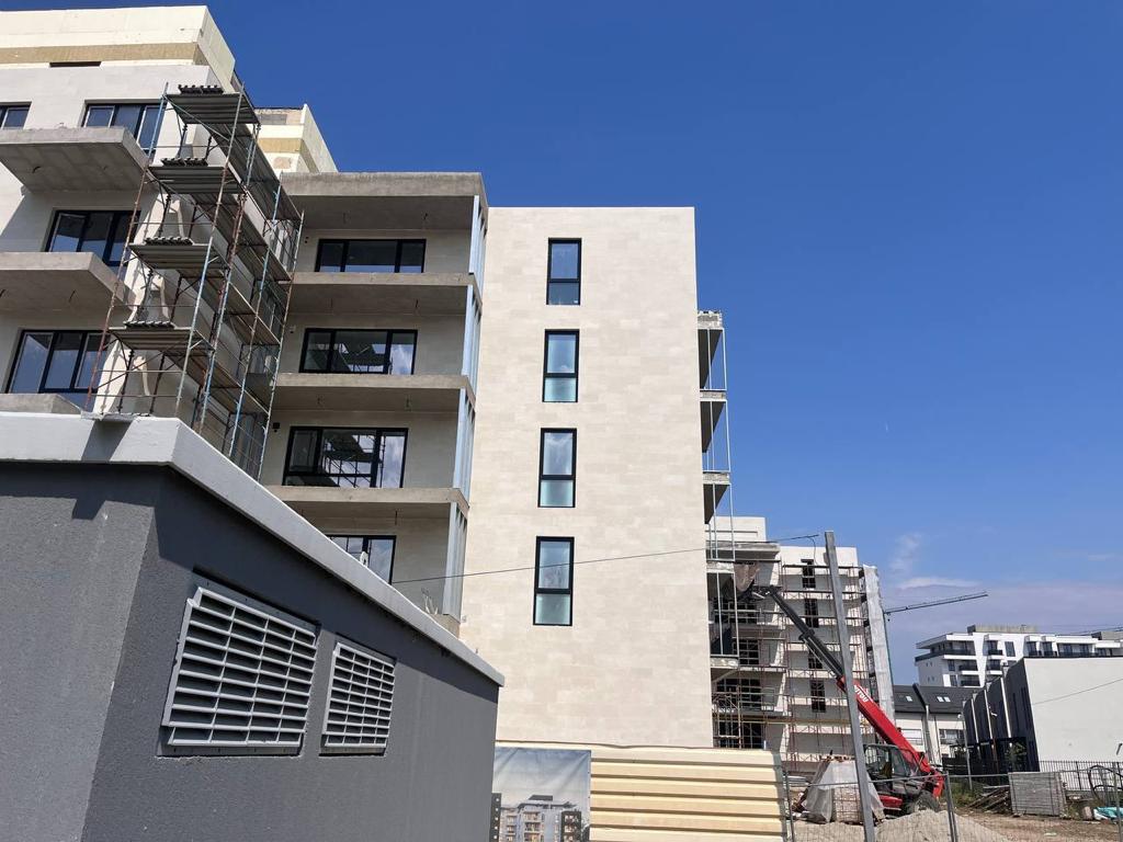 Corner of a block in beige flexible stone with a grey service building in front and a tower crane behind