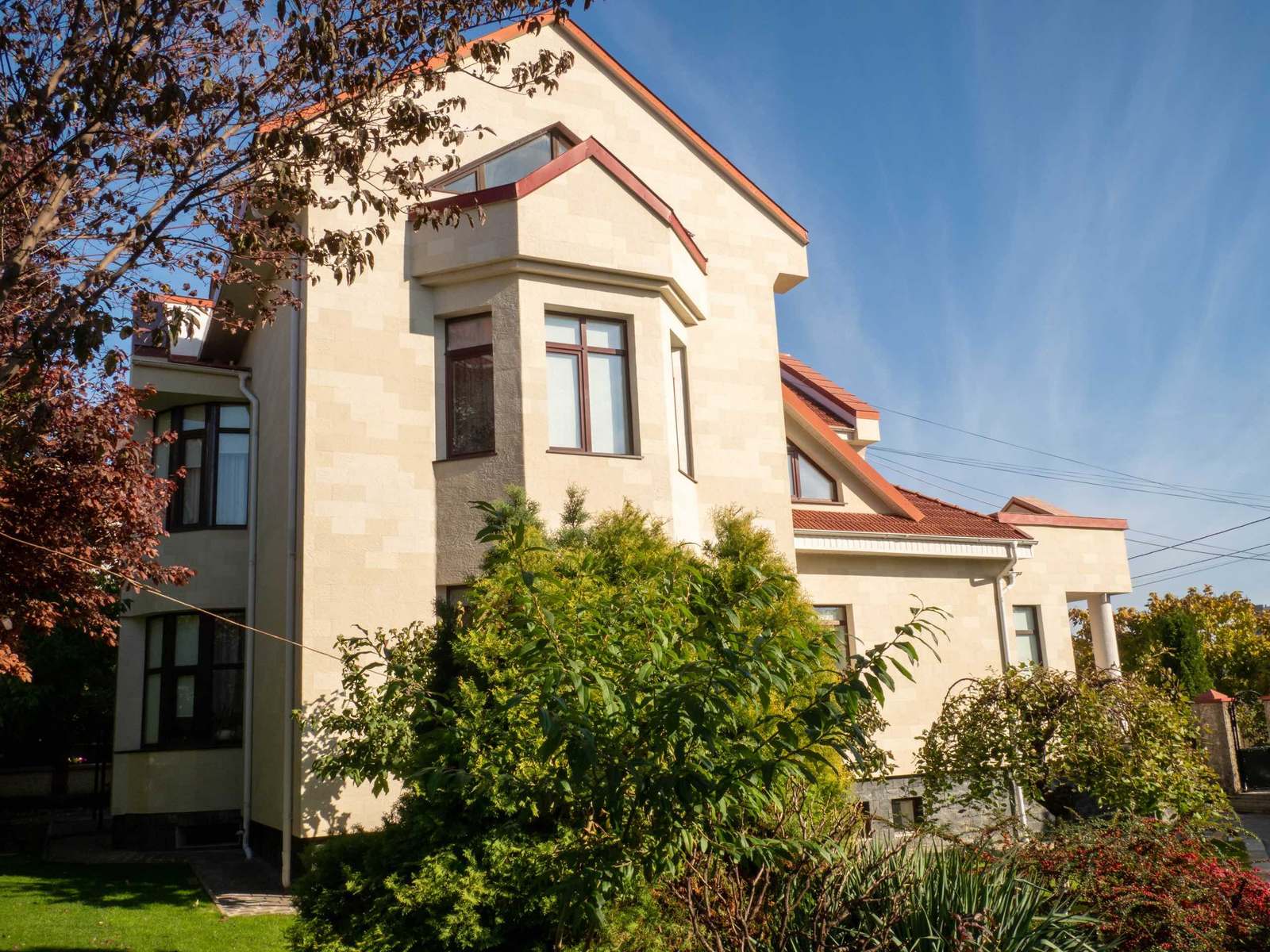 The house from the garden, with bay window, tall gable and beige flexible stone facade among shrubs