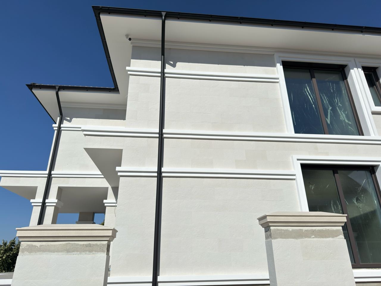 House corner from the garden side, balcony, white cornices and a black downpipe on flexible stone